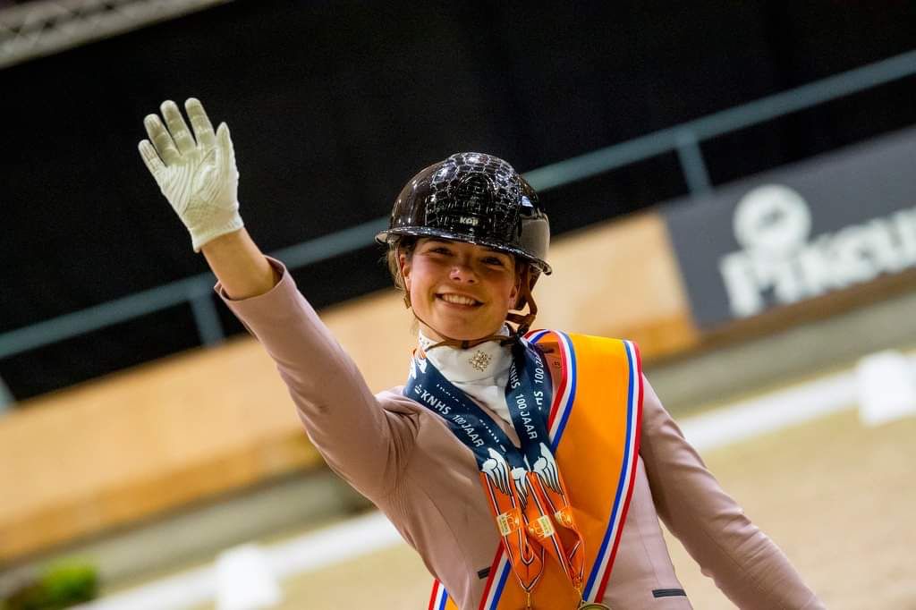 Febe van Zwambagt waving to the camera during the prize-giving of her Dutch National Championship title, wearing a JuulC competition jacket.